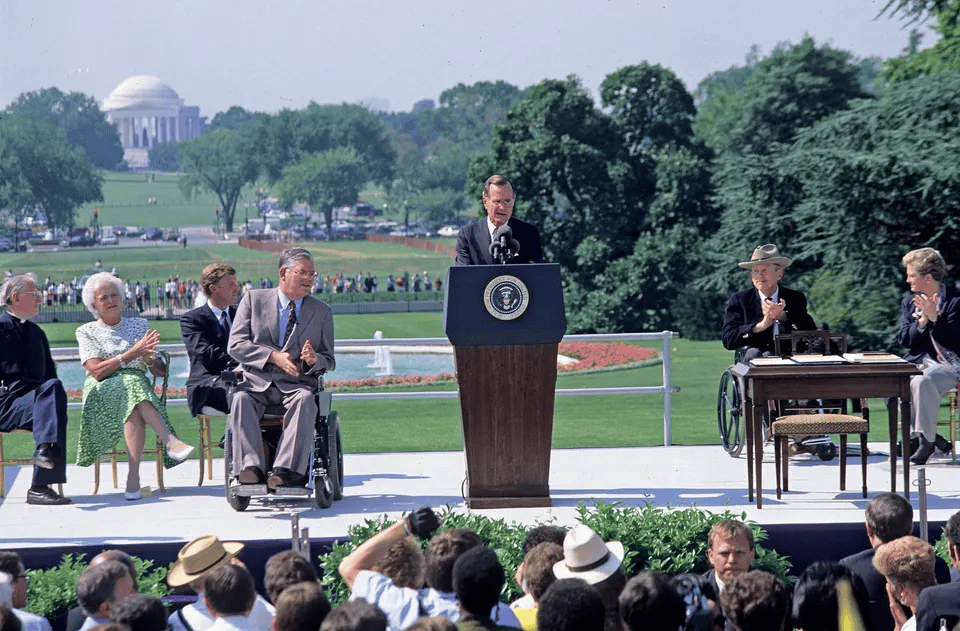 Man in suit on stage in from of a podium. He has two people in wheel chairs next to him wearing suits. Behind them is a grassy field with trees