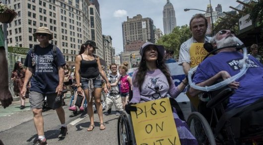 People participate in the first annual Disability Pride Parade. Two wheelchair users in front and dozens of people marching behind.