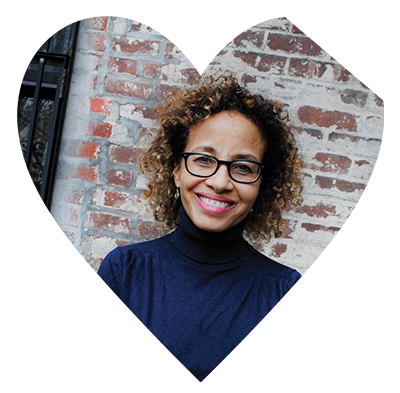 Heart with top left corner cut off. Inside a women with glasses, wearing a blue turtleneck standing in front of brick wall