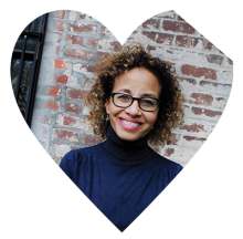 Heart with top left corner cut off. Inside a women with glasses, wearing a blue turtleneck standing in front of brick wall