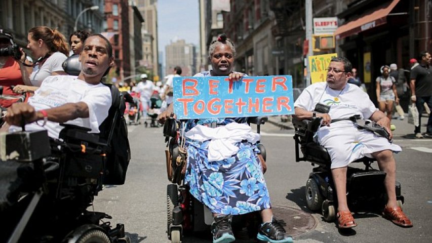 Three people in wheelchairs marching in the NYC Disability Pride Parade. One holding a sign saying 'Better Together'