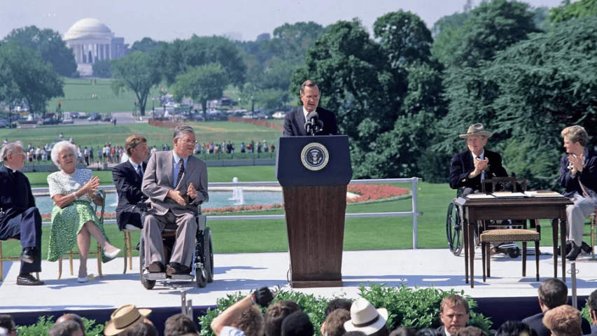 Man in suit on stage in from of a podium. He has two people in wheel chairs next to him wearing suits. Behind them is a grassy field with trees