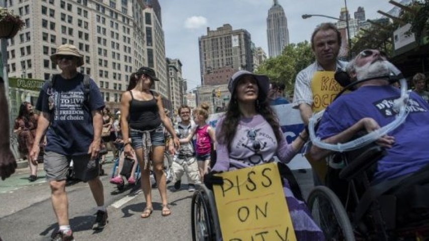 People participate in the first annual Disability Pride Parade. Two wheelchair users in front and dozens of people marching behind.